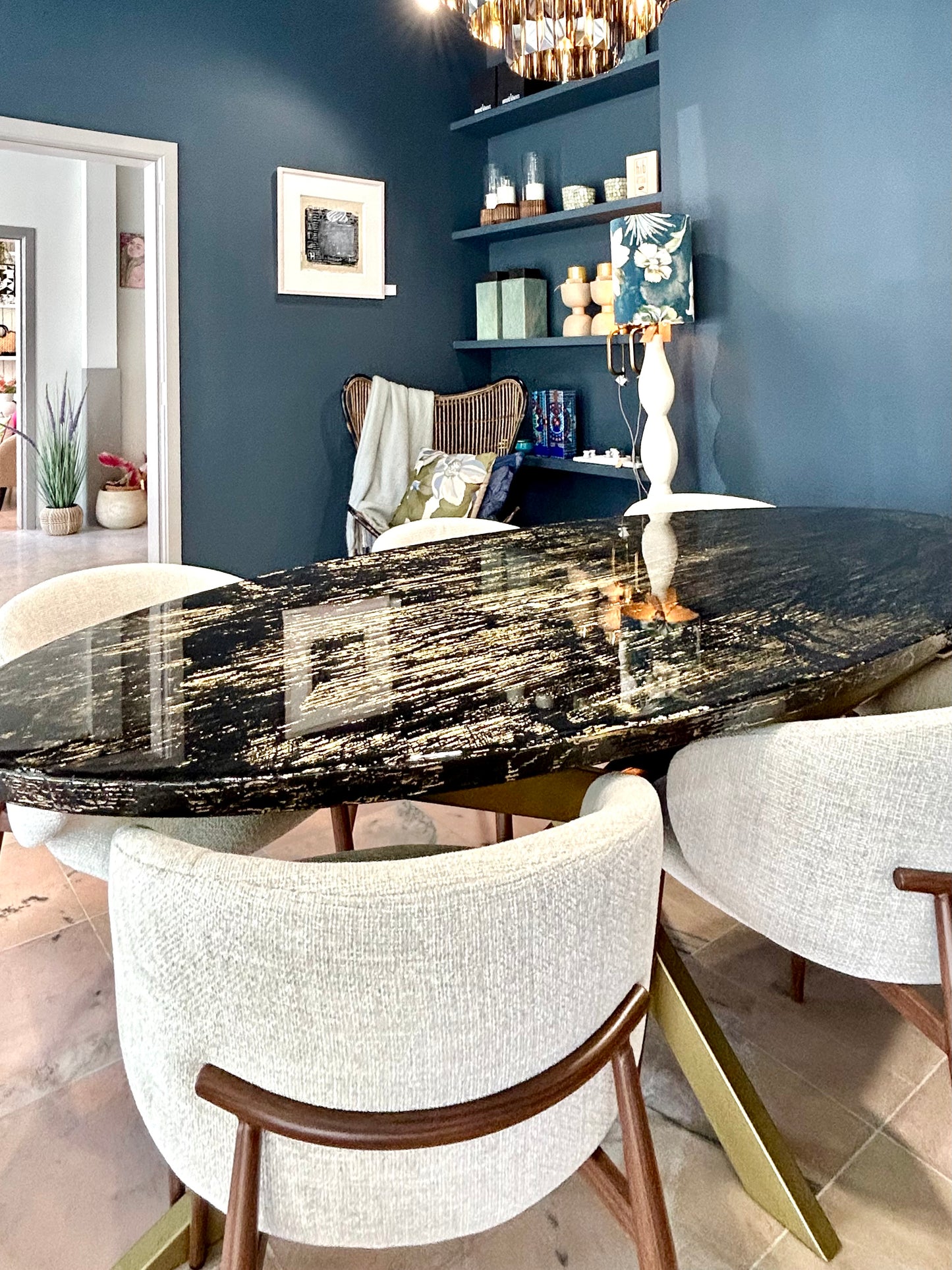 Een ovale eettafel met een glanzend zwart blad met goudkleurige nerven staat centraal in een modern ingerichte ruimte. Rond de tafel staan zes lichtbeige, gestoffeerde stoelen met houten poten en een halfronde rugleuning. Aan de muur hangt een donkerblauwe 3D-wandbekleding met organische patronen. Boven de tafel hangt een grote glazen designlamp en links staat een console met decoratieve objecten en een moderne tafellamp.
