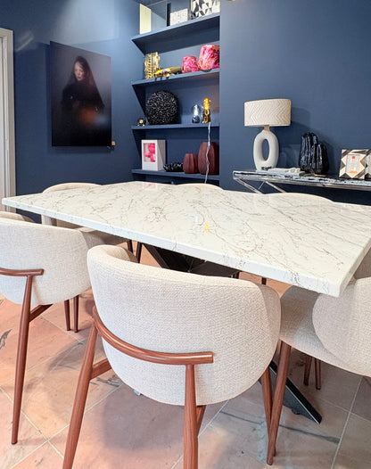Luxe eettafel met een glanzend, rechthoekig blad in marmerlook met beige en gouden en zilveren aders. Rondom de tafel staan vier gestoffeerde eetstoelen met houten poten en afgeronde rugleuningen. De tafel staat in een moderne showroom met een donkere, structuurwand, een design hanglamp