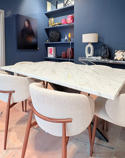 Luxe eettafel met een glanzend, rechthoekig blad in marmerlook met beige en gouden en zilveren aders. Rondom de tafel staan vier gestoffeerde eetstoelen met houten poten en afgeronde rugleuningen. De tafel staat in een moderne showroom met een donkere, structuurwand, een design hanglamp