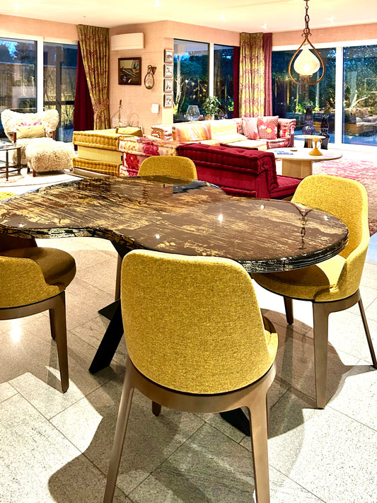 Organisch gevormde eettafel met een hoogglanzend donker blad vol goudachtige nerven en structuren. De tafel staat in een warme, luxe ingerichte woonkamer met veel kleur: gele stoffen stoelen rondom de tafel, een rood-roze zithoek op de achtergrond, grote ramen met uitzicht op een tuin en sfeervolle verlichting aan het plafond.