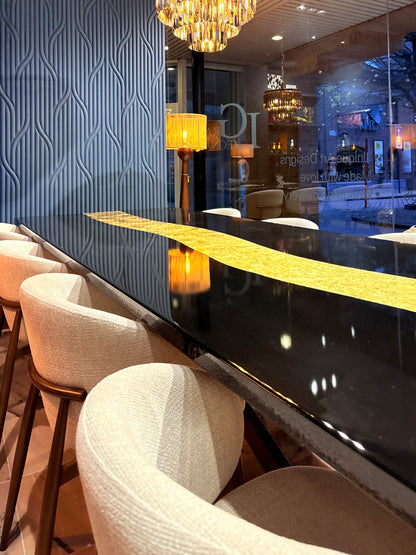 Langwerpige, glanzende zwarte eettafel met een opvallende goudkleurige, organische strook in het midden. Rondom de tafel staan crèmekleurige, modern vormgegeven stoelen met houten poten. Op de achtergrond zijn grote ramen te zien met uitzicht op een straat, warme verlichting van hanglampen en een muur met een blauw 3D-patroon.