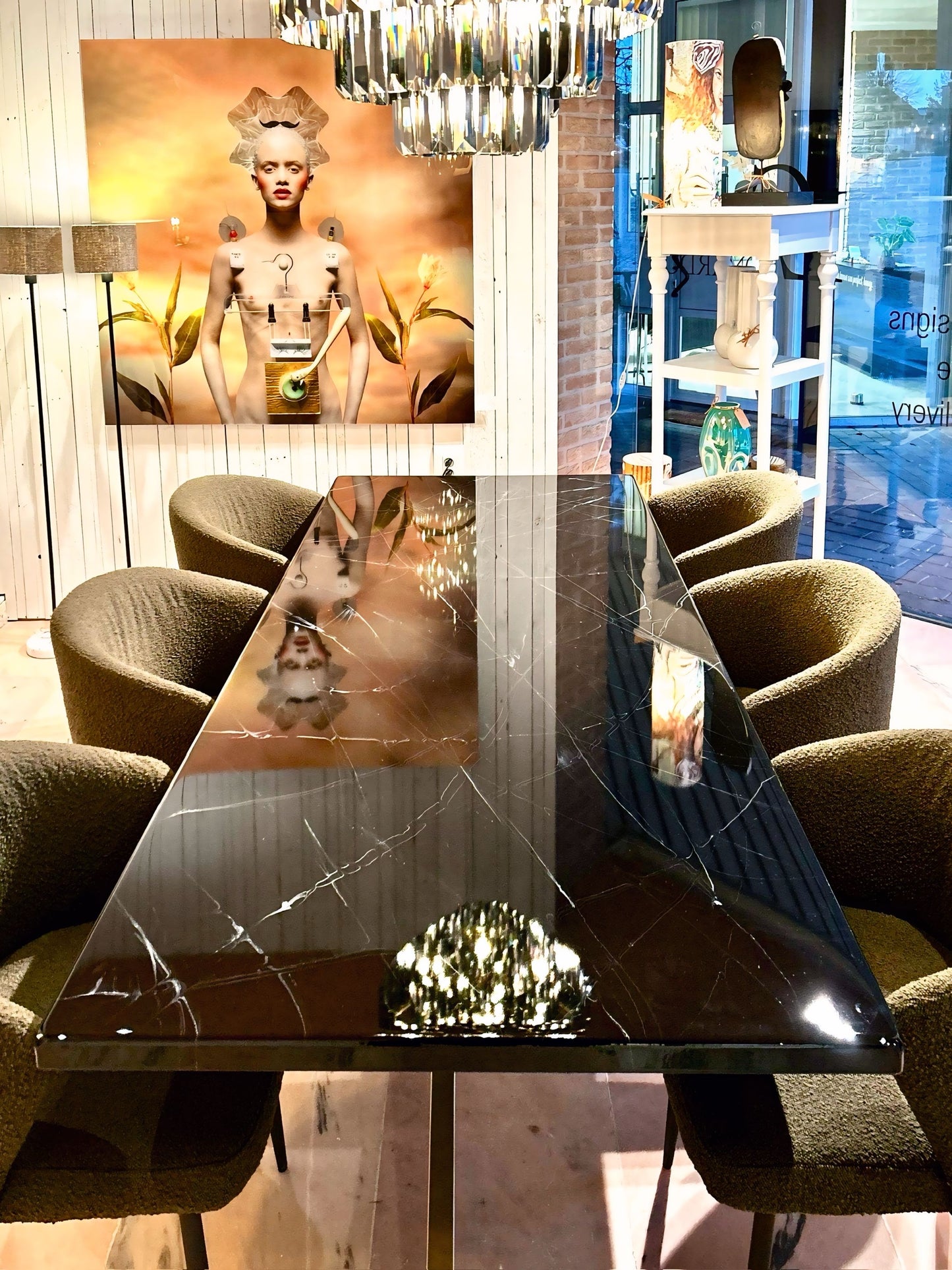 Rechthoekige eettafel met een glanzend zwart marmeren tafelblad met witte aders, omringd door olijfgroene gestoffeerde stoelen. Boven de tafel hangt een moderne chandelier die weerspiegelt in het blad. Op de achtergrond staat een grote kunstfoto van een stijlvol gestileerde vrouw tegen een warmgele achtergrond, in een sfeervol ingerichte ruimte met houten wand en decoratieve items.