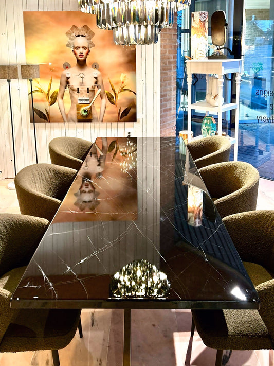 Rechthoekige eettafel met een glanzend zwart marmeren tafelblad met witte aders, omringd door olijfgroene gestoffeerde stoelen. Boven de tafel hangt een moderne chandelier die weerspiegelt in het blad. Op de achtergrond staat een grote kunstfoto van een stijlvol gestileerde vrouw tegen een warmgele achtergrond, in een sfeervol ingerichte ruimte met houten wand en decoratieve items.