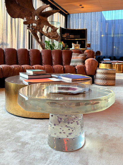 Een moderne woonkamer met een laag, transparant epoxy bijzettafeltje dat is gevuld met paarse mineralen in de voet en een geode-achtige steen in het midden van het blad. Het tafeltje staat op een zacht, licht tapijt. Op de achtergrond staat een ronde, goudkleurige salontafel met boeken erop, een grote cognackleurige lederen bank en een opvallend decoratief dinosaurus-schedelobject dat boven de bank hangt. De ruimte heeft hoge ramen met doorschijnende gordijnen en een warme, luxe sfeer