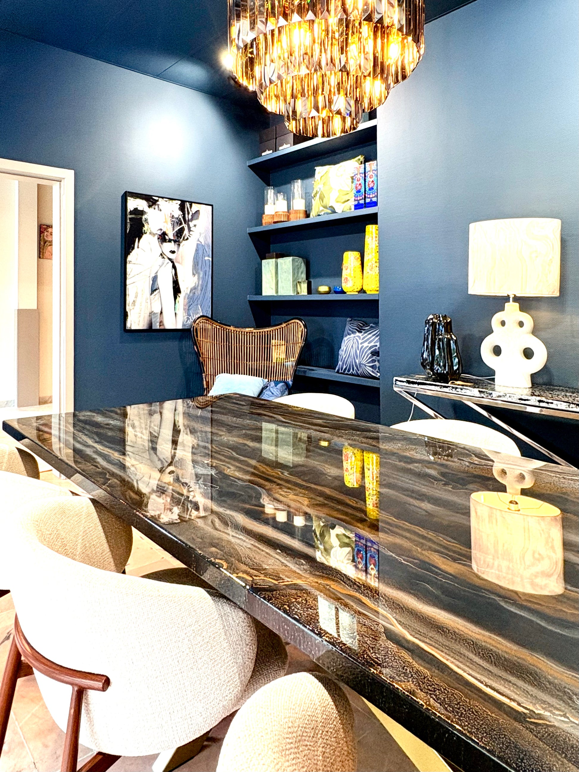 Luxe eetkamer met een grote rechthoekige tafel met een glanzend epoxyblad in zwarte, gouden en grijze marmerachtige patronen. Rondom de tafel staan lichtbeige, gestoffeerde armstoelen met houten details. Aan de muur hangen twee ronde, goudkleurige wandlampen en boven de tafel hangt een moderne chandelier met veel glinsterende elementen