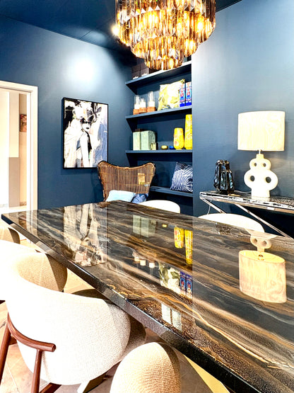 Luxe eetkamer met een grote rechthoekige tafel met een glanzend epoxyblad in zwarte, gouden en grijze marmerachtige patronen. Rondom de tafel staan lichtbeige, gestoffeerde armstoelen met houten details. Aan de muur hangen twee ronde, goudkleurige wandlampen en boven de tafel hangt een moderne chandelier met veel glinsterende elementen