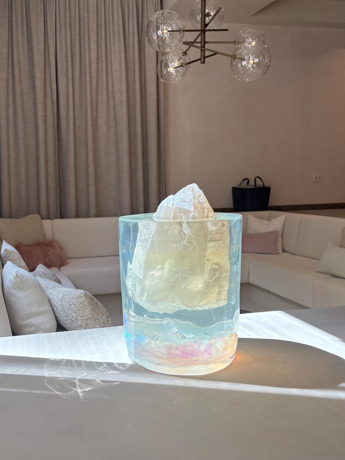 Een kunstzinnig sculptuur van epoxyhars met een edelsteen in het midden staat op een witte tafel in een moderne woonkamer. Het transparante, iriserende epoxyobject vangt het zonlicht, waardoor kleurrijke lichtreflecties op het tafelblad ontstaan. Achter het kunstwerk staan een witte hoekbank met kussens, beige gordijnen en een moderne lamp met glazen bollen.