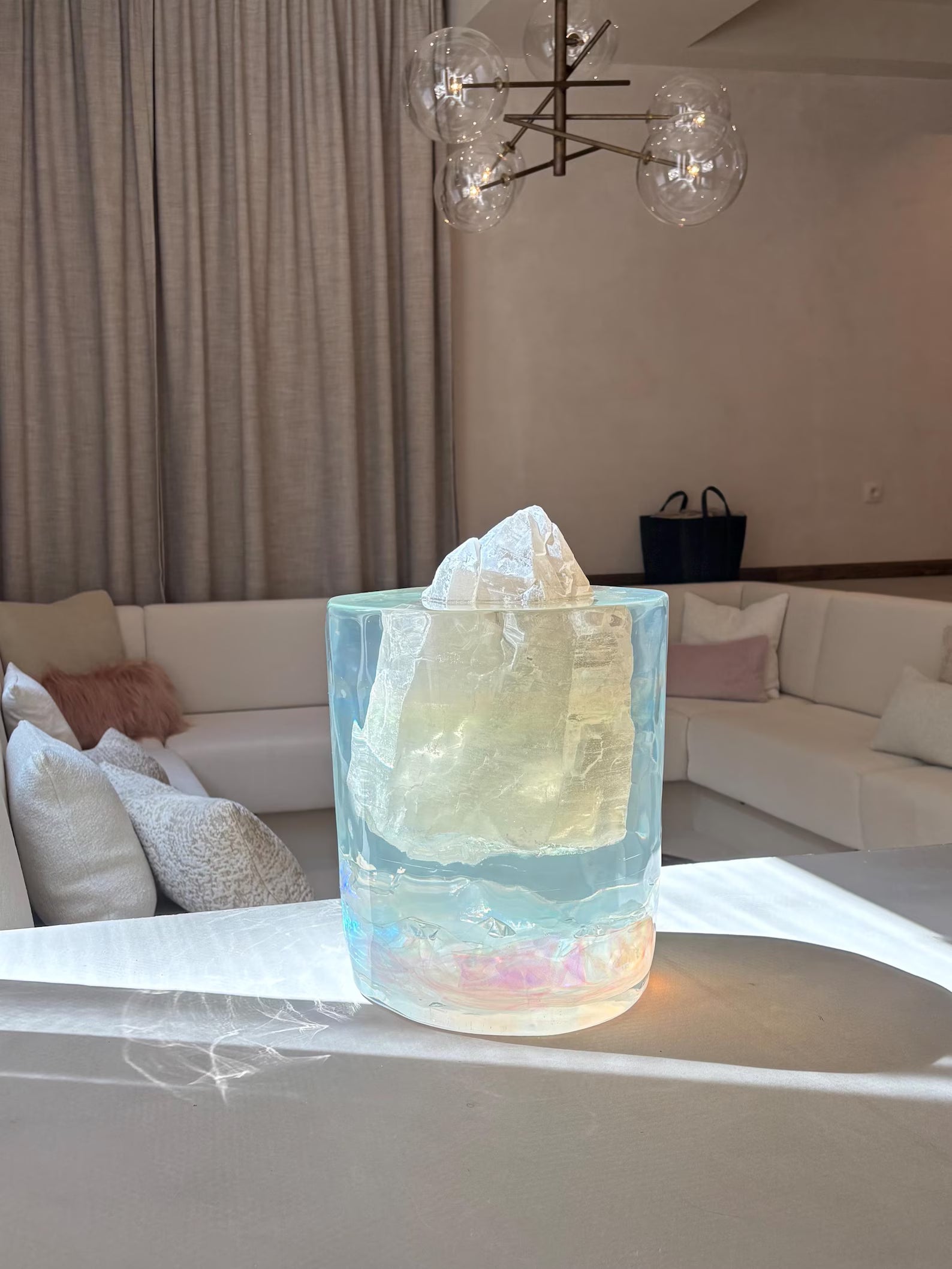 Een kunstzinnig sculptuur van epoxyhars met een edelsteen in het midden staat op een witte tafel in een moderne woonkamer. Het transparante, iriserende epoxyobject vangt het zonlicht, waardoor kleurrijke lichtreflecties op het tafelblad ontstaan. Achter het kunstwerk staan een witte hoekbank met kussens, beige gordijnen en een moderne lamp met glazen bollen.